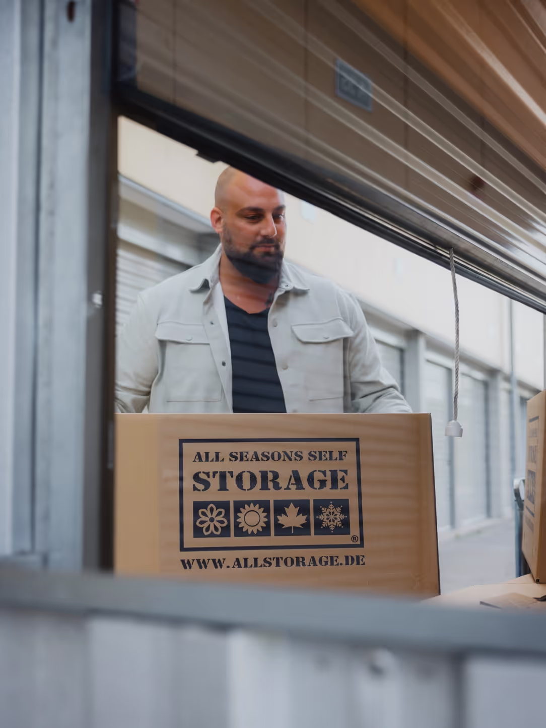 Ein Mann mit Glatze und Bart steht hinter einem großen Karton und schaut darauf. Der Karton ist mit dem Logo und der Aufschrift ‘ALL SEASONS SELF STORAGE’ sowie den Symbolen für die vier Jahreszeiten (Blume, Sonne, Ahornblatt und Schneeflocke) bedruckt. Die Website ‘www.allstorage.de’ ist ebenfalls auf dem Karton zu sehen. Der Mann trägt eine helle Jacke und ein dunkles T-Shirt. Im Hintergrund sind Lagerräume mit geöffneten Rolltoren zu erkennen.
