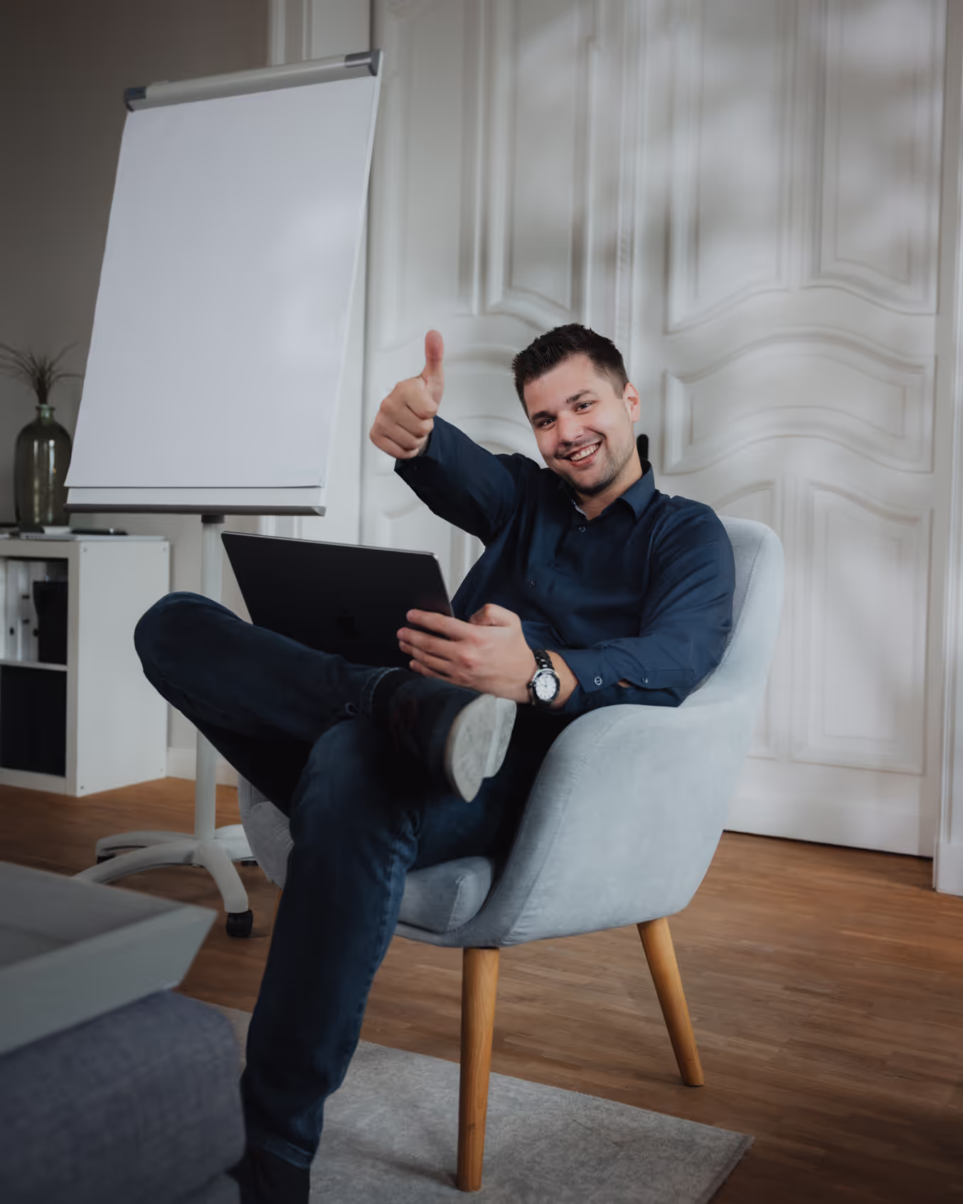 Ein Mann in einem dunklen Hemd sitzt an einem Tisch und blickt freundlich in die Kamera. Seine entspannte Körperhaltung und das warme Lächeln vermitteln eine einladende und sympathische Ausstrahlung. Im Hintergrund sind unscharfe Elemente zu sehen, die den Fokus auf die Person lenken. Die Beleuchtung ist weich und hebt sein Gesicht angenehm hervor.