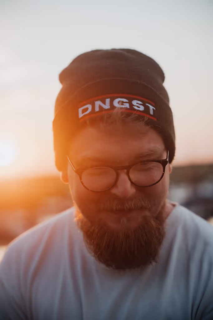 Ein Mann sitzt entspannt auf einem Sofa vor einem dunkel gehaltenen Hintergrund. Er trägt ein schlichtes T-Shirt und schaut leicht zur Seite, als wäre er in Gedanken vertieft. Die Beleuchtung ist weich und hebt sein Gesicht subtil hervor, wodurch eine ruhige und reflektierte Atmosphäre entsteht.