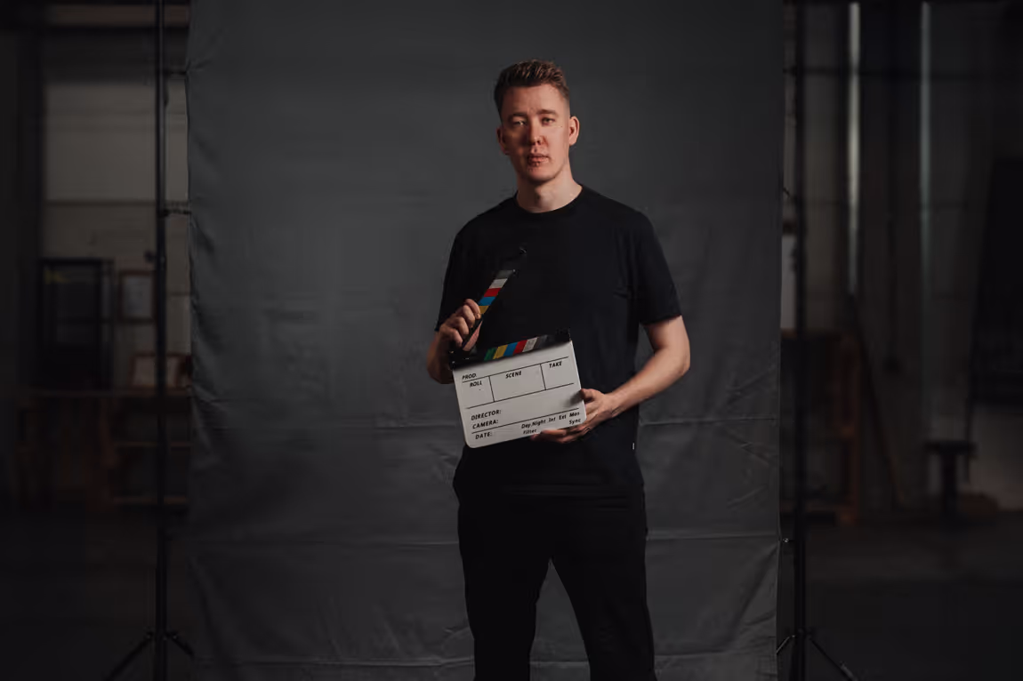 Ein Mann in schwarzer Kleidung steht vor einem neutralen grauen Hintergrund und hält eine Filmklappe. Er blickt ruhig und fokussiert in die Kamera. Der Hintergrund ist schlicht, mit leichter Beleuchtung, während im Randbereich unscharfe Studioelemente wie Stative und Möbel zu erkennen sind. Die Szene vermittelt einen professionellen und kreativen Eindruck.