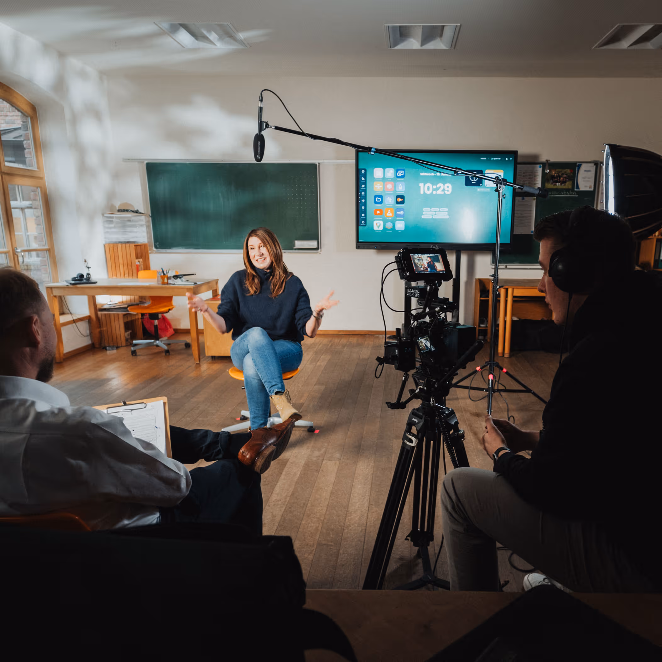 Eine Frau sitzt auf einem Stuhl vor einer Kamera und spricht, während zwei Männer sie im Klassenzimmer mit Film- und Tonausrüstung aufnehmen.